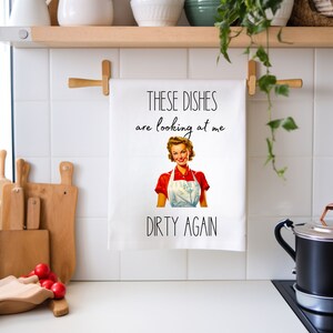 May include: White kitchen towel with a retro illustration of a woman in a red shirt and white apron. The text on the towel reads "These dishes are looking at me dirty again."