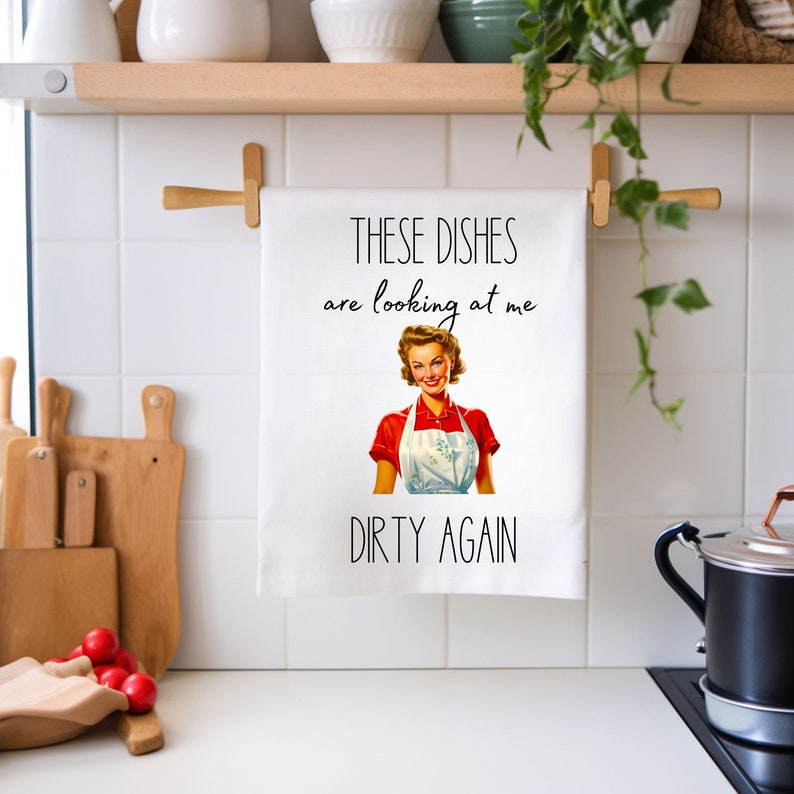 May include: White kitchen towel with a retro illustration of a woman in a red shirt and white apron. The text on the towel reads "These dishes are looking at me dirty again."