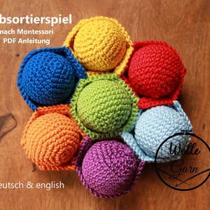 May include: A rainbow-colored crocheted ball sorting game for children. The balls are arranged in a circle and are made of different colors, including red, orange, yellow, green, blue, and purple. The game is labeled "Farbsortierspiel nach Montessori PDF Anleitung" and "deutsch & english".