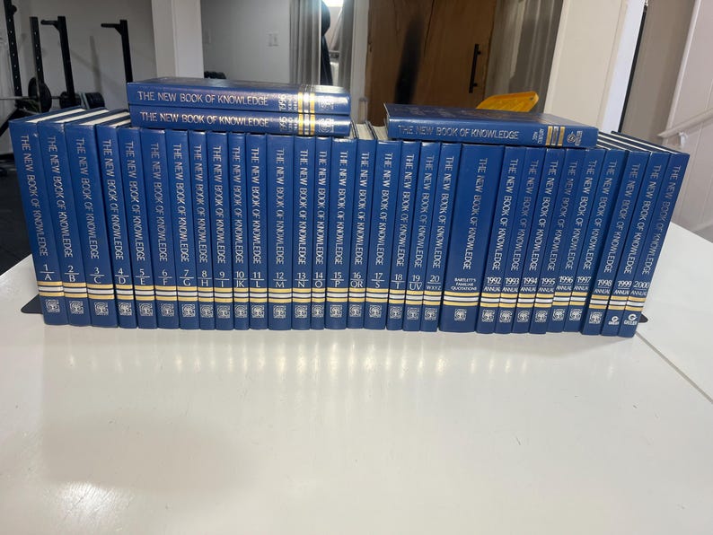 Może przedstawiać: Kolekcja niebieskich książek w twardej oprawie zatytułowanych "The New Book of Knowledge". Grzbiety mają złote litery i symbole. Książki ułożone są na białej powierzchni, niekt&oacute;re ułożone jeden na drugim. Kolekcja obejmuje tomy z lat 1992-2000.