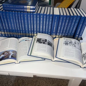 Może przedstawiać: Kolekcja niebieskich książek w twardej oprawie zatytułowanych "The New Book of Knowledge" jest prezentowana na białej powierzchni. Kilka książek jest otwartych, ukazując ilustrowane strony. Książki są ułożone w rzędach, sugerując zestaw referencyjny.