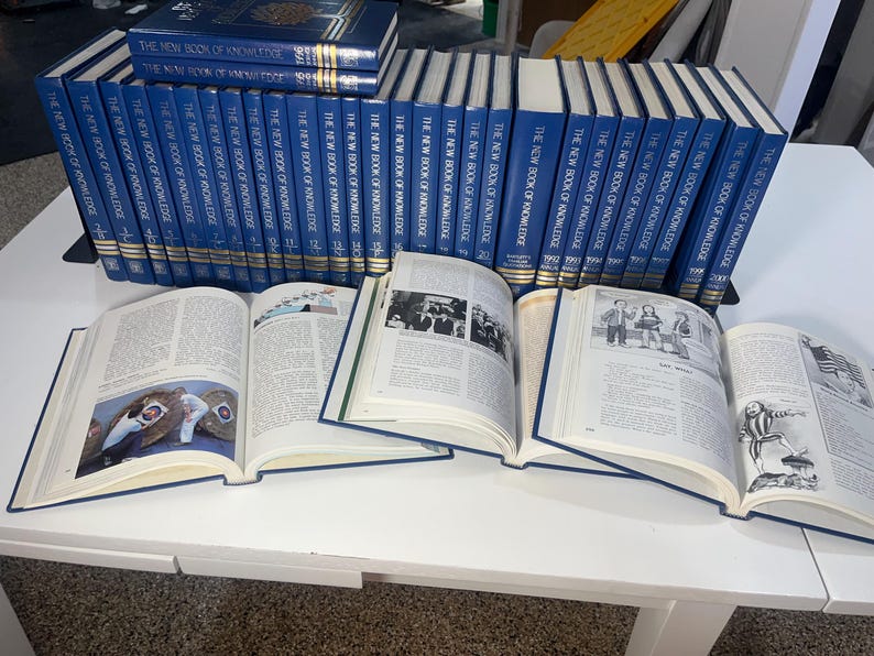 Może przedstawiać: Kolekcja niebieskich książek w twardej oprawie zatytułowanych "The New Book of Knowledge" jest prezentowana na białej powierzchni. Kilka książek jest otwartych, ukazując ilustrowane strony. Książki są ułożone w rzędach, sugerując zestaw referencyjny.