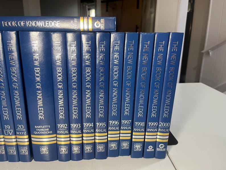 Może przedstawiać: Kolekcja niebieskich książek w twardej oprawie zatytułowanych "The New Book of Knowledge" i "Book of Knowledge". Grzbiety pokazują lata 1992-2000 oraz "Bartlett's Familiar Quotations". Książki są ułożone na p&oacute;łce.