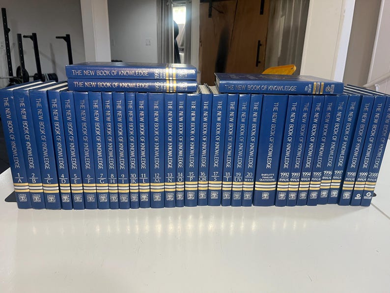 Może przedstawiać: Kolekcja niebieskich książek w twardej oprawie zatytułowanych "The New Book of Knowledge". Grzbiety prezentują złote litery i cyfry, tomy ułożone są na białej powierzchni. Zestaw zawiera tomy z lat 1992-2000.