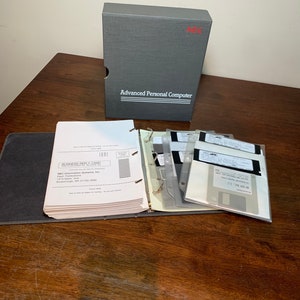 May include: A gray box with the text "NEC Advanced Personal Computer" on the front. The box is open and there are several floppy disks inside. The disks are in a binder with a business reply card.