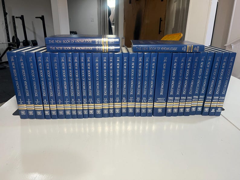 Może przedstawiać: Kolekcja niebieskich książek w twardej oprawie zatytułowanych "The New Book of Knowledge". Grzbiety mają złote litery i numerację. Książki ułożone są na białej powierzchni, niekt&oacute;re ułożone w stos.