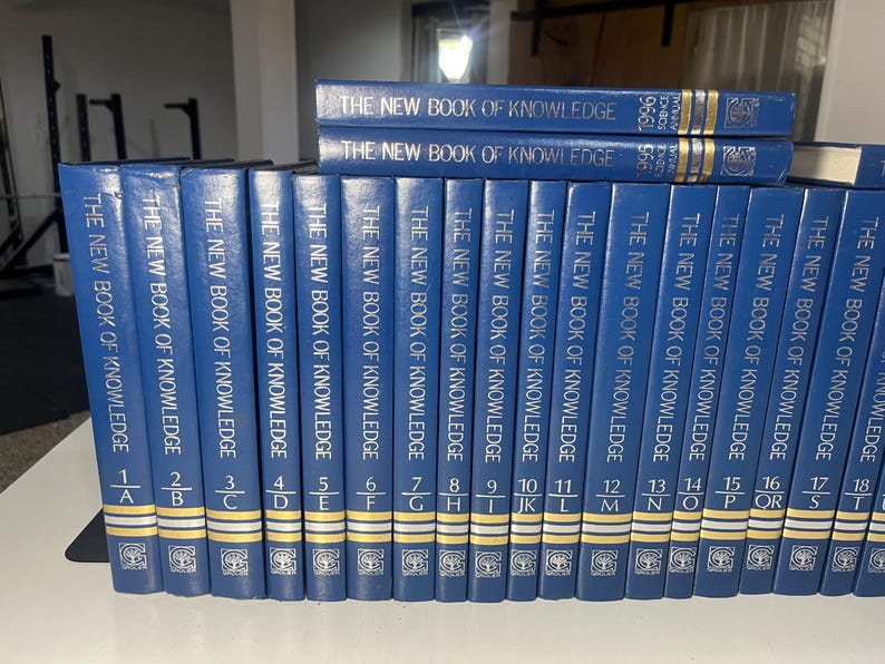 Może przedstawiać: Kolekcja niebieskich książek w twardej oprawie zatytułowanych "The New Book of Knowledge". Grzbiety są niebieskie ze złotymi literami i numeracją. Książki ułożone są w rzędzie, z kilkoma dodatkowymi tomami ułożonymi na wierzchu.