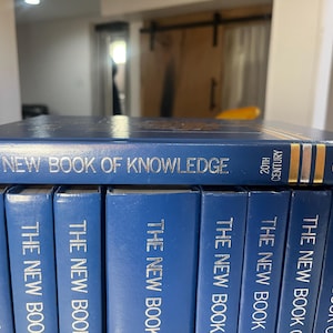 Może przedstawiać: Kolekcja niebieskich książek w twardej oprawie zatytułowanych "The New Book of Knowledge". G&oacute;rna książka wyświetla tytuł srebrnymi literami, z napisem "20th Century" i logo Grolier w kolorze złotym. Książki są starannie ułożone.