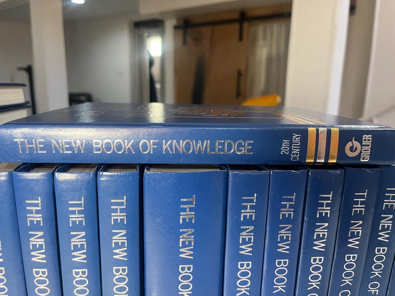 Może przedstawiać: Kolekcja niebieskich książek w twardej oprawie zatytułowanych "The New Book of Knowledge". G&oacute;rna książka wyświetla tytuł srebrnymi literami, z napisem "20th Century" i logo Grolier w kolorze złotym. Książki są starannie ułożone.