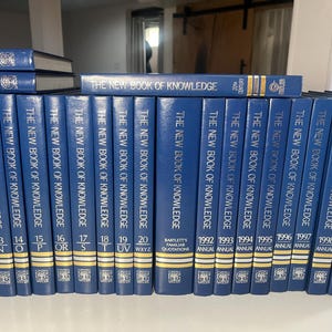 Może przedstawiać: Kolekcja niebieskich książek w twardej oprawie zatytułowanych "The New Book of Knowledge". Grzbiety pokazują tytuł i numery tom&oacute;w, w tym wydania roczne od 1992 do 2000 roku. Książki są ułożone na p&oacute;łce.