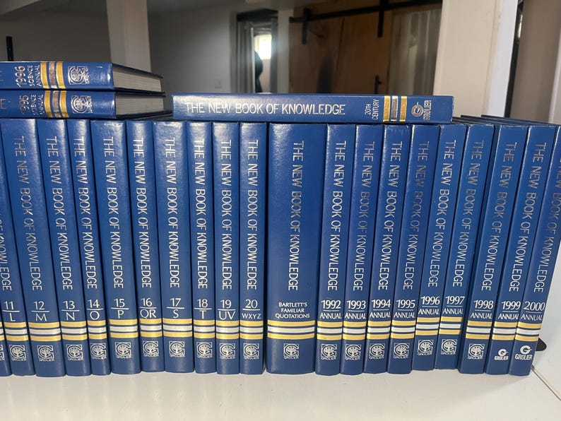 Może przedstawiać: Kolekcja niebieskich książek w twardej oprawie zatytułowanych "The New Book of Knowledge". Grzbiety pokazują tytuł i numery tom&oacute;w, w tym wydania roczne od 1992 do 2000 roku. Książki są ułożone na p&oacute;łce.