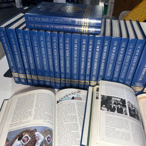 Może przedstawiać: Kolekcja niebieskich książek w twardej oprawie zatytułowanych "The New Book of Knowledge". Kilka książek jest ułożonych w stos, inne w rzędzie. Otwarte książki ukazują zadrukowane strony z ilustracjami i tekstem.