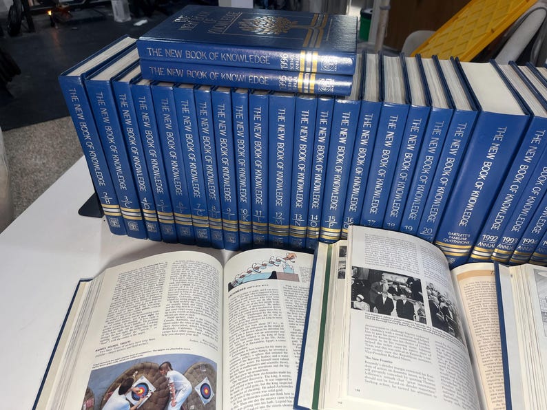 Może przedstawiać: Kolekcja niebieskich książek w twardej oprawie zatytułowanych "The New Book of Knowledge". Kilka książek jest ułożonych w stos, inne w rzędzie. Otwarte książki ukazują zadrukowane strony z ilustracjami i tekstem.