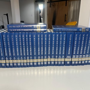 Może przedstawiać: Kolekcja niebieskich książek w twardej oprawie zatytułowanych "The New Book of Knowledge" ułożonych na białej powierzchni. Grzbiety mają złote litery i numery, niekt&oacute;re tomy są ułożone w stos.