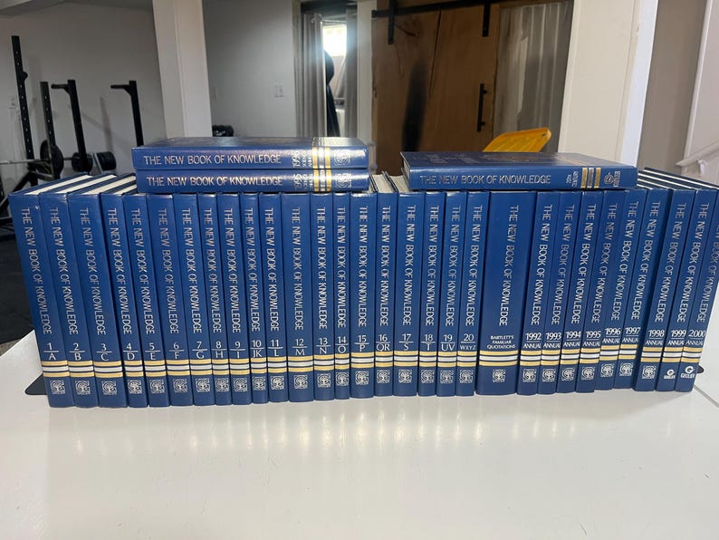 Może przedstawiać: Kolekcja niebieskich książek w twardej oprawie zatytułowanych "The New Book of Knowledge" ułożonych na białej powierzchni. Grzbiety mają złote litery i numery, niekt&oacute;re tomy są ułożone w stos.