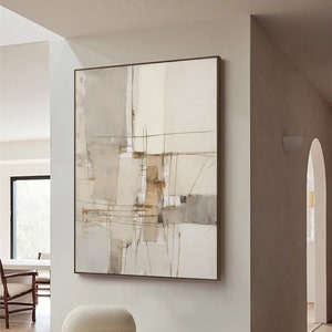 Puede incluir: Pintura abstracta en tonos beige, marrón y blanco con finas líneas negras. La pintura está enmarcada y colgada en una pared blanca sobre una silla blanca mullida y un taburete de madera.
