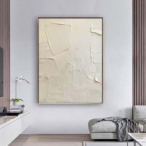 May include: A white abstract painting with a textured, layered effect. The painting is framed in a thin gold frame and hangs on a white wall in a modern living room.