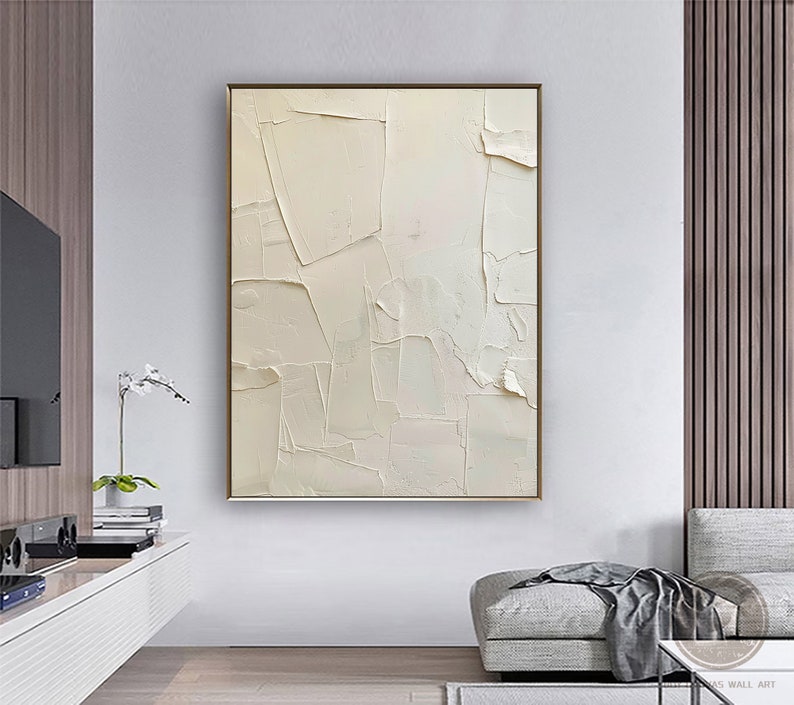 May include: A white abstract painting with a textured, layered effect. The painting is framed in a thin gold frame and hangs on a white wall in a modern living room.