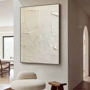 May include: A white abstract painting with a textured surface hangs on a white wall above a white fluffy chair and a wooden stool.