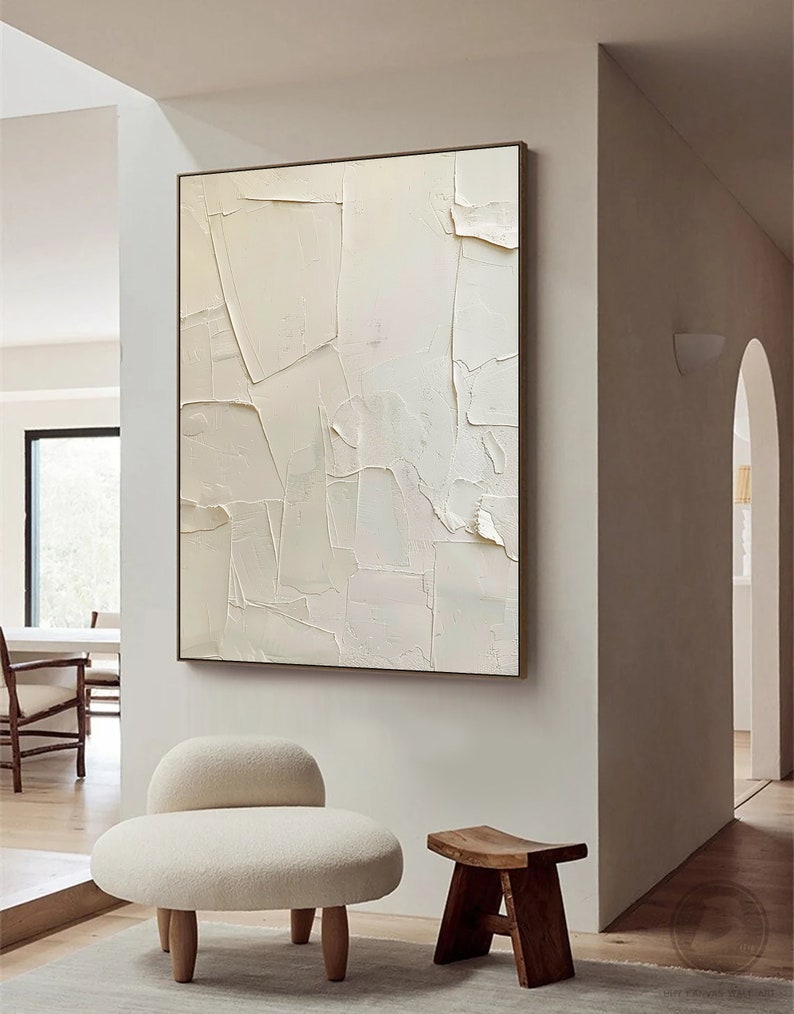 May include: A white abstract painting with a textured surface hangs on a white wall above a white fluffy chair and a wooden stool.