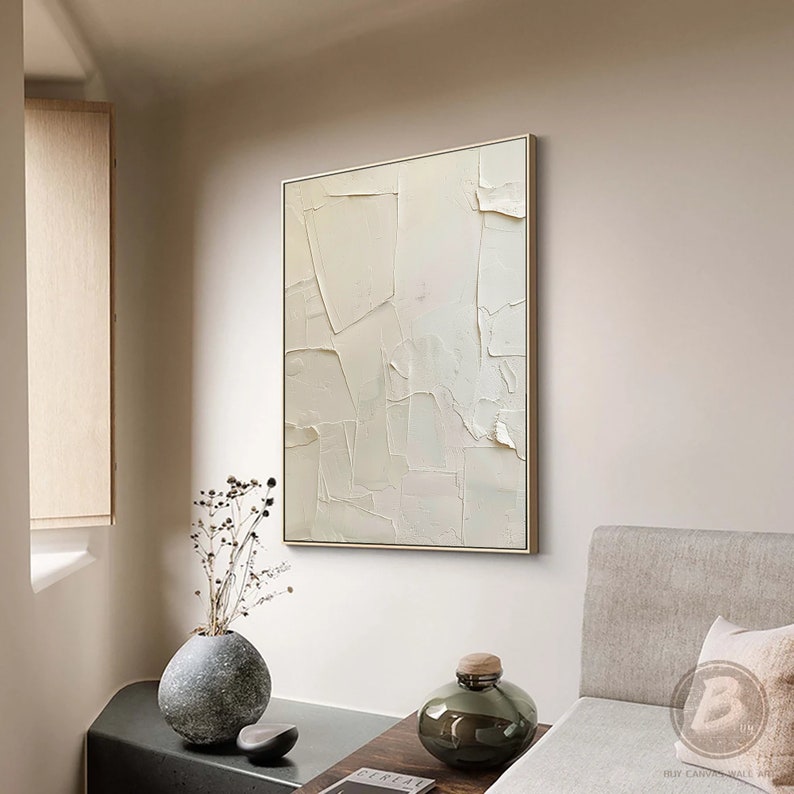 May include: A white abstract painting with a textured surface, framed in a gold frame, hanging on a white wall. The painting is in a modern living room setting with a couch and a vase of dried flowers.