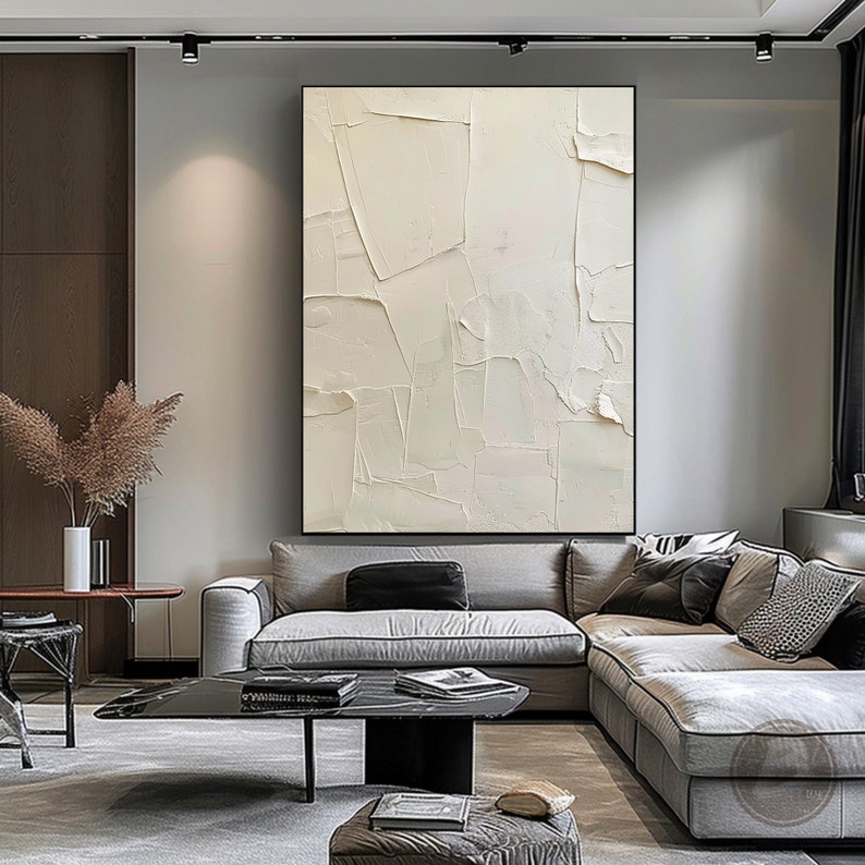 May include: A white abstract painting with a textured surface. The painting is hanging on a wall in a living room with a gray sectional sofa and a black coffee table.