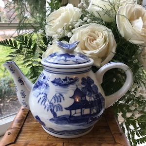 May include: White and blue ceramic teapot with a butterfly-shaped lid finial. The teapot features a scenic blue and white design with pagodas and trees. The teapot is set against a backdrop of white roses and greenery.