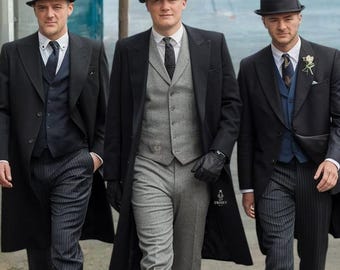 May include: Three men wearing black suits and hats walk down a street. The man in the middle is wearing a gray vest and gray pants. The man on the left is wearing a black vest and black pants. The man on the right is wearing a blue vest and black pants. The men are all wearing black shoes.