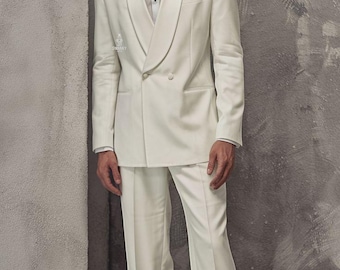 Op de afbeelding: Een crèmekleurig dubbelknoops pak met een sjaalkraag, gecombineerd met een wit overhemd. Het pak is van Swanky Suits. Het model draagt een wit pak en witte schoenen. De achtergrond is een getextureerde grijze muur.