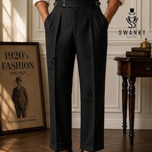 May include: Dark charcoal gray trousers featuring a high-waisted design with double pleats, a wide waistband with side adjusters, and cuffed hems. The fabric has a textured appearance, and the garment is paired with a white shirt and black loafers.