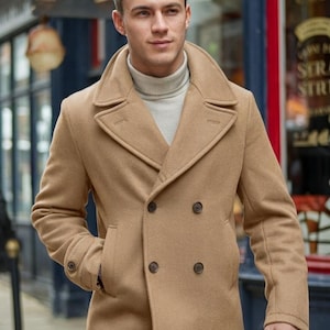 May include: A tan double-breasted pea coat with a notched collar and two rows of dark brown buttons. The coat has two front pockets and a matching tan color. The person is wearing a light gray turtleneck.