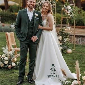 Puede incluir: Una pareja posa al aire libre, el hombre con un traje verde oscuro y la mujer con un vestido de novia blanco. El traje tiene chaqueta, chaleco y pantalón a juego. El vestido tiene una larga cola. Un arco floral está en el fondo.