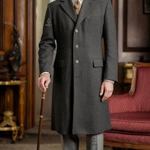 May include: A charcoal gray overcoat with a matching hat and gray trousers. The coat has a classic three-button closure and a notched lapel. The person is holding a wooden cane. The brand logo "Swanky Suits" is visible.