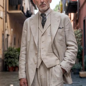 Puede incluir: Un hombre con un traje de lino beige claro con una corbata estampada. El traje tiene un diseño de tres piezas con un chaleco y una chaqueta. La chaqueta tiene un bolsillo de parche en el lado izquierdo. El hombre está de pie en un callejón estrecho.