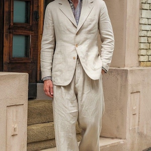 May include: A light beige linen suit with a straw boater hat. The suit includes a two-button blazer and wide-leg trousers. The hat has a navy blue band. The outfit is completed with beige loafers. The logo "SWANKY SUITS" is visible.