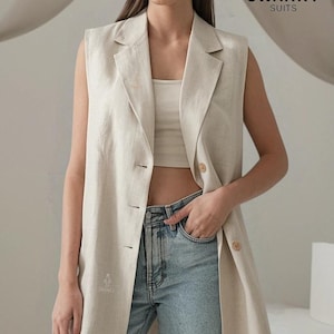 Könnte beinhalten: Eine lange, hellbeige Leinenweste mit einem V-Ausschnitt und Knopfverschluss. Die Weste hat eine lockere Passform und wird über einem weißen Tanktop und einer hellblauen Jeans getragen.