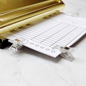 Peut inclure: Une feuille de papier blanche avec un motif quadrillé et le mot "ACCOMPLISH" imprimé dessus. La feuille est maintenue en place par deux pinces à lier en plastique transparent. La feuille est partiellement insérée dans un perforateur de papier de couleur or.
