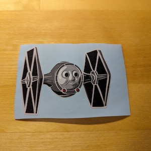 May include: A black and white TIE fighter with a cartoon face on the front. The TIE fighter is on a light blue background.