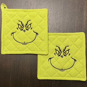 May include: Two lime green quilted pot holders with a black embroidered Grinch face. The Grinch's eyes are yellow. One pot holder has a loop for hanging. The pot holders are square.