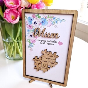 May include: Wooden framed picture with the word "Mum" in a cursive font, floral design, and a puzzle piece design with names. The text reads "The piece that holds us all together". A single puzzle piece is on the surface.