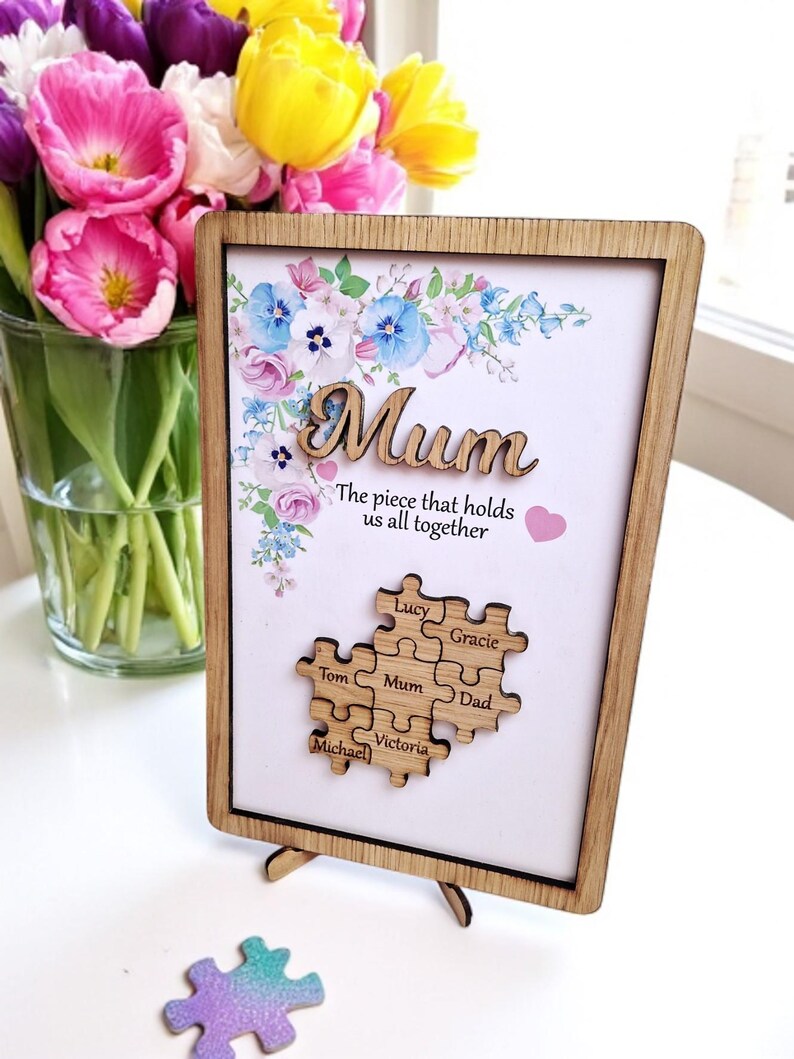 May include: Wooden framed picture with the word "Mum" in a cursive font, floral design, and a puzzle piece design with names. The text reads "The piece that holds us all together". A single puzzle piece is on the surface.