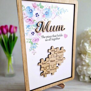 May include: Wooden framed artwork with the word "Mum" in a cursive font, floral accents, and a puzzle piece design. The text "The piece that holds us all together" is below the word "Mum". The puzzle pieces have names on them.