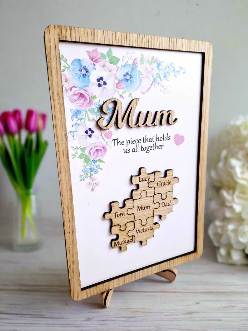 May include: Wooden framed artwork with the word "Mum" in a cursive font, floral accents, and a puzzle piece design. The text "The piece that holds us all together" is below the word "Mum". The puzzle pieces have names on them.