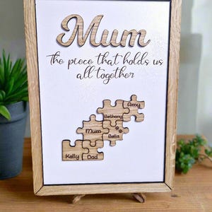 May include: Wooden framed artwork with the word "Mum" in cursive script. The text "the piece that holds us all together" is below. Puzzle pieces with names "Kelly", "Dad", "Mum", "Bethany", "Bella", and "Amy" are displayed.
