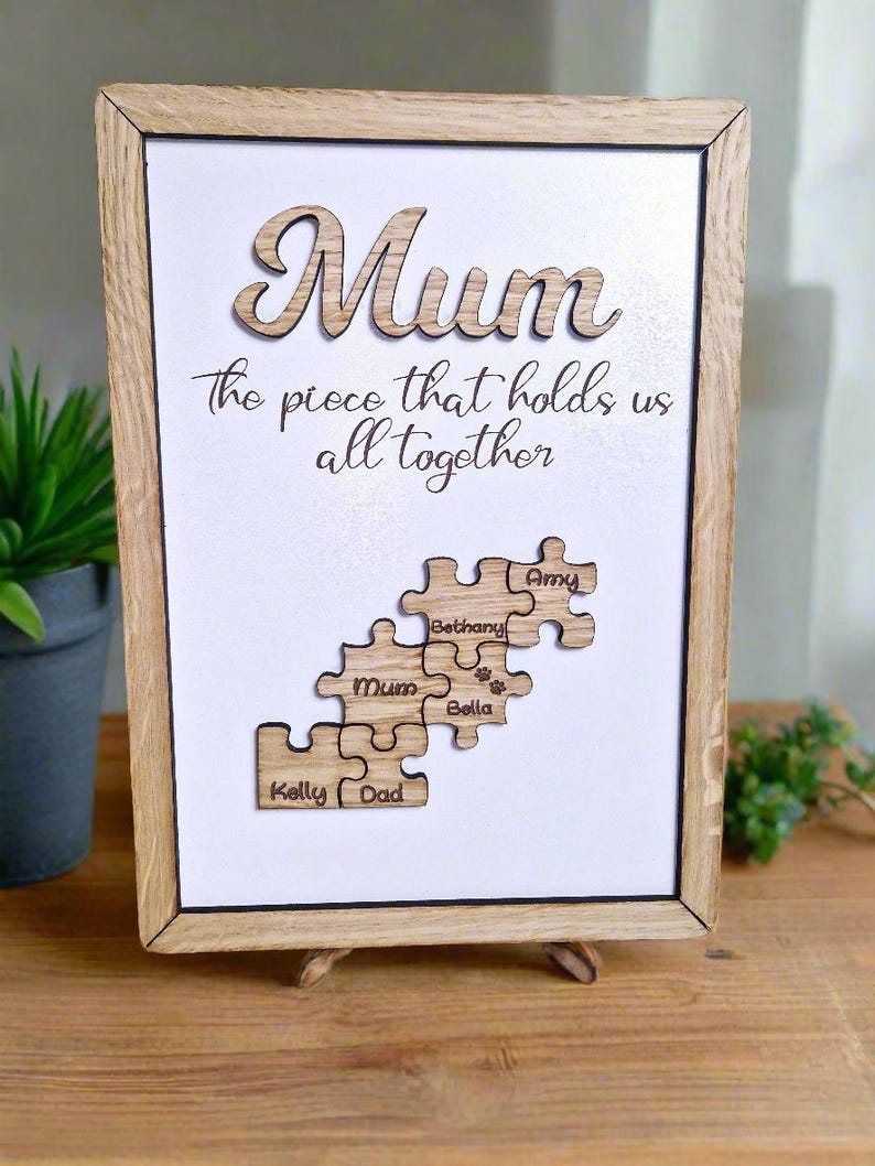 May include: Wooden framed artwork with the word "Mum" in cursive script. The text "the piece that holds us all together" is below. Puzzle pieces with names "Kelly", "Dad", "Mum", "Bethany", "Bella", and "Amy" are displayed.
