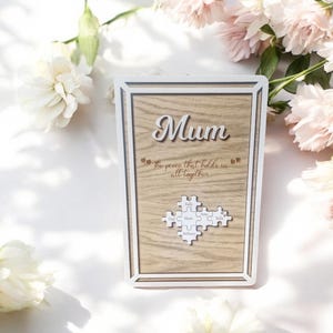 May include: Wooden framed plaque with the word "Mum" in cursive. The plaque has a light wood grain background and includes the text "the pieces that hold us all together". A puzzle piece design is at the bottom.