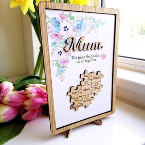 May include: Wooden framed sign with the word "Mum" and the text "The piece that holds us all together." Floral design with a puzzle piece with names. The frame is supported by a wooden stand.