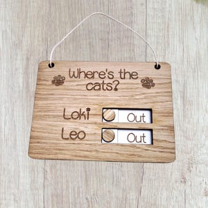 May include: A wooden sign with the text "Where's the cats?" and paw prints. It features sliders for "Loki" and "Leo" with "Out" indicators. The sign is made of light brown wood and has a string for hanging.