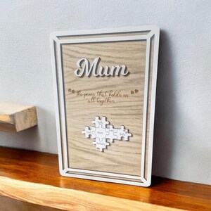 May include: A wooden framed sign with the word "Mum" in cursive. The sign includes the phrase "the piece that holds us all together" and a puzzle piece design with names. The frame is white, and the background is a light wood grain.