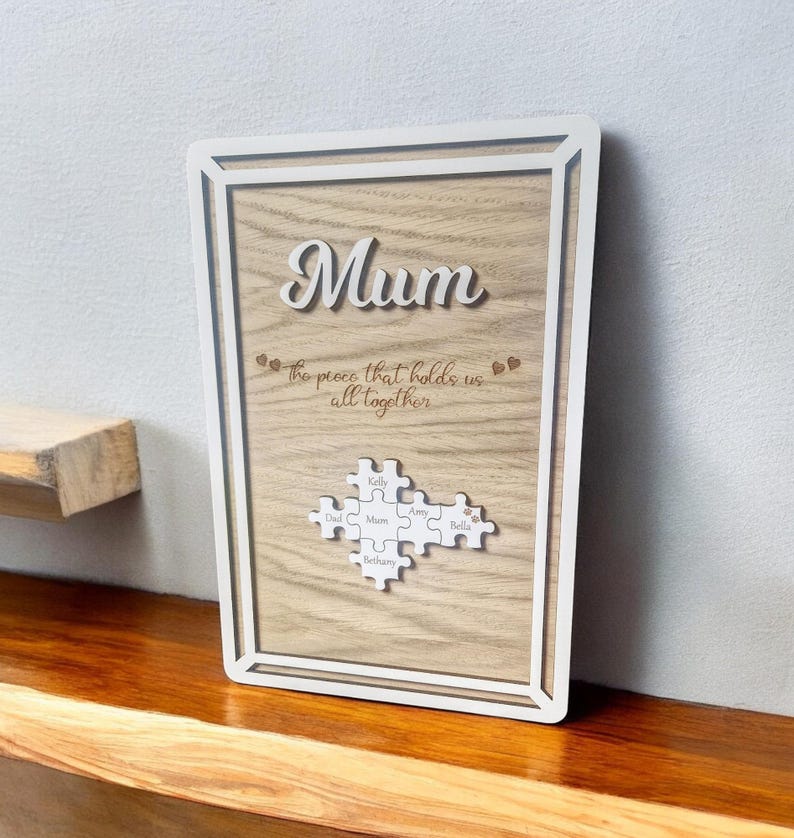 May include: A wooden framed sign with the word "Mum" in cursive. The sign includes the phrase "the piece that holds us all together" and a puzzle piece design with names. The frame is white, and the background is a light wood grain.